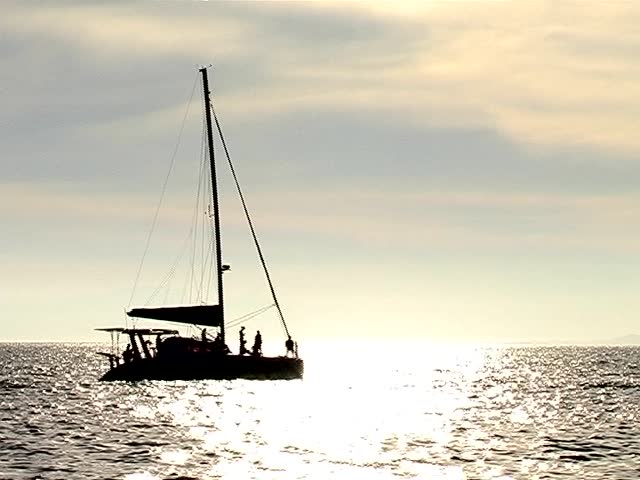 Catamaran sailing away on the ocean, through the sunset as a silhouette 