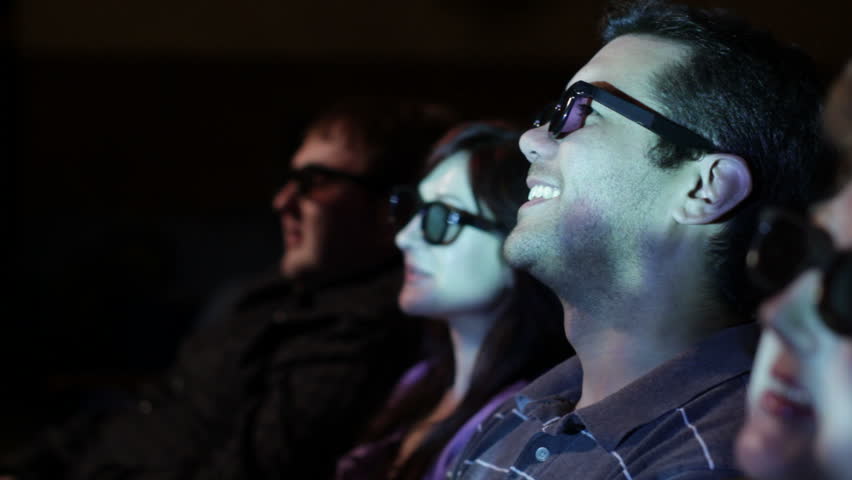 Young man watching a 3D movie with a group of friends, exchanging comments. Focus on him with a small dolly move and projections on his face.