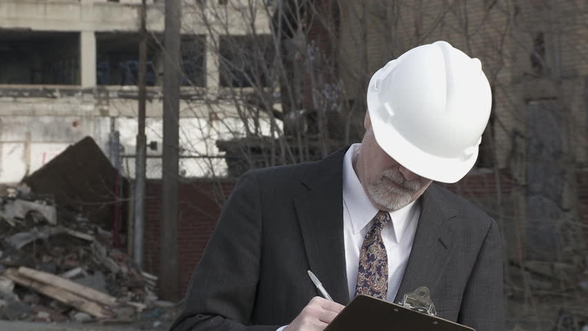 Developer (could be an architect, investor, contractor or other business person) walks into shot and looks around at the collapsed buildings around him, making notes. Close up.