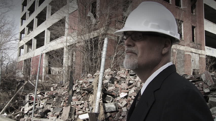 Developer walks into shot and looks around at the collapsed buildings around him, making notes. Big close up on developer, hand held camera.
