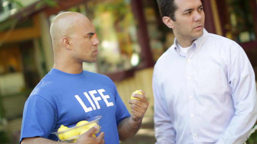 Man in a blue T-shirt that says "LIFE" sells a lemon to a shady character on the street.