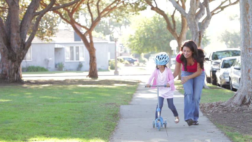 Proud mother watches as daughter rides on scooter towards camera. Shot on Canon 5d Mk2 with a frame rate of 30fps