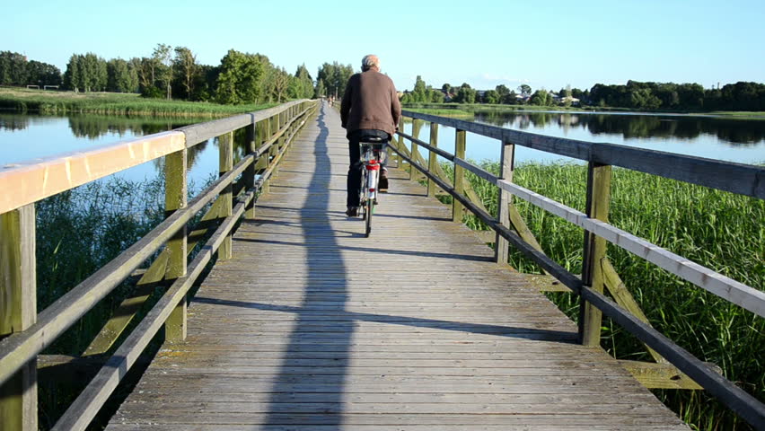 grey head man drive bicicle on wooden long bridge through lake.