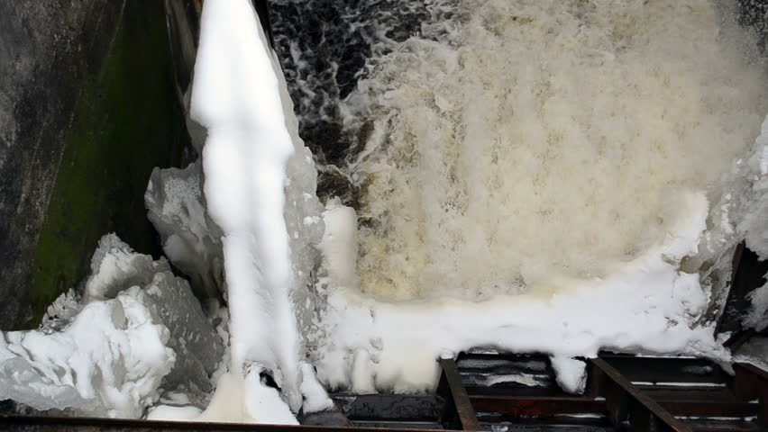 strong water current flow with splash spray in old vintage dam frozen with ice and snow in winter.