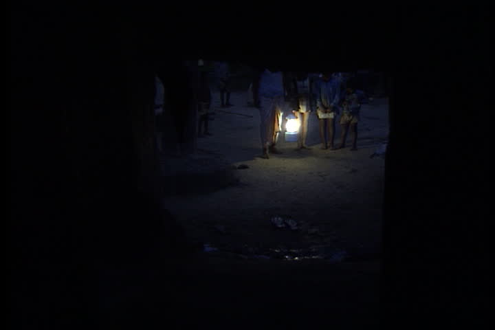 SULTANPUR, INDIA - JULY 28, 1999: Man enters dark hut carrying lit solar lantern. Family gathers around light and start to read.