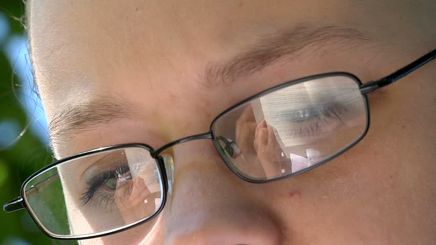 A teenage girl reads a book under a tree - reflected in her glasses