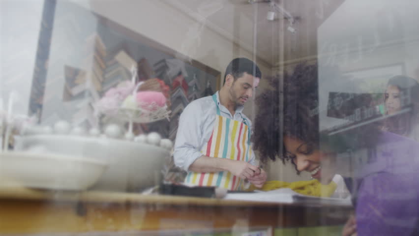 A friendly male shopkeeper is at work behind the counter of a quaint village shop and serving a female customer. As seen from outside looking in, through the storefront window. In slow motion.