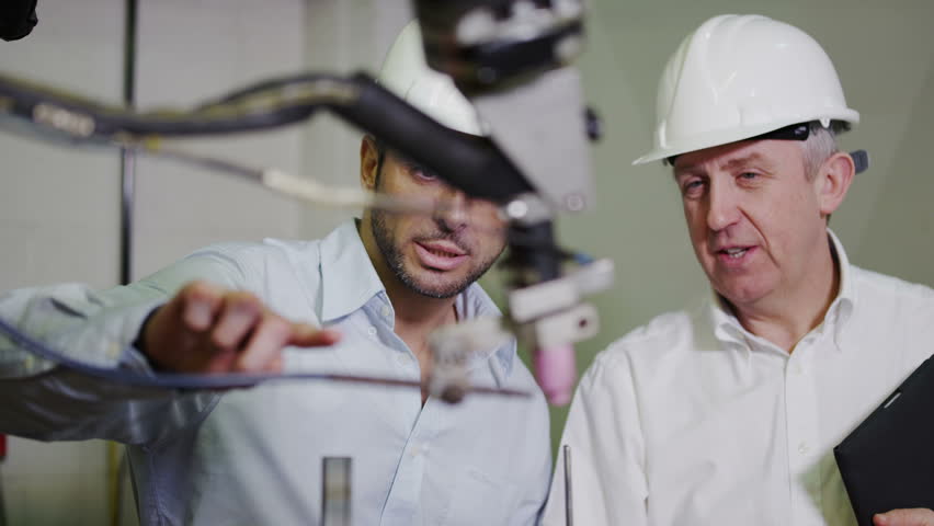 A mature male factory manager is with a young male client or colleague and is explaining how some of the plant machinery works. 