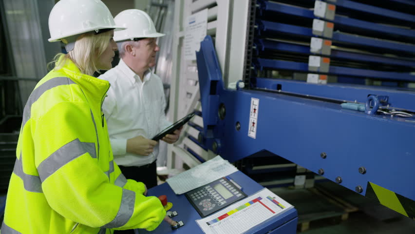 A female worker within a factory or warehouse is being shown by her foreman how to operate some heavy machinery.
