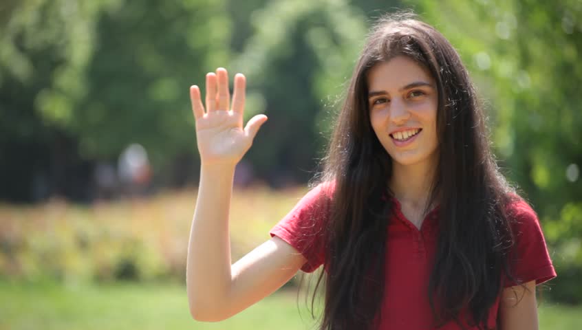 Brunette young adult woman waves her hand to the camera.
