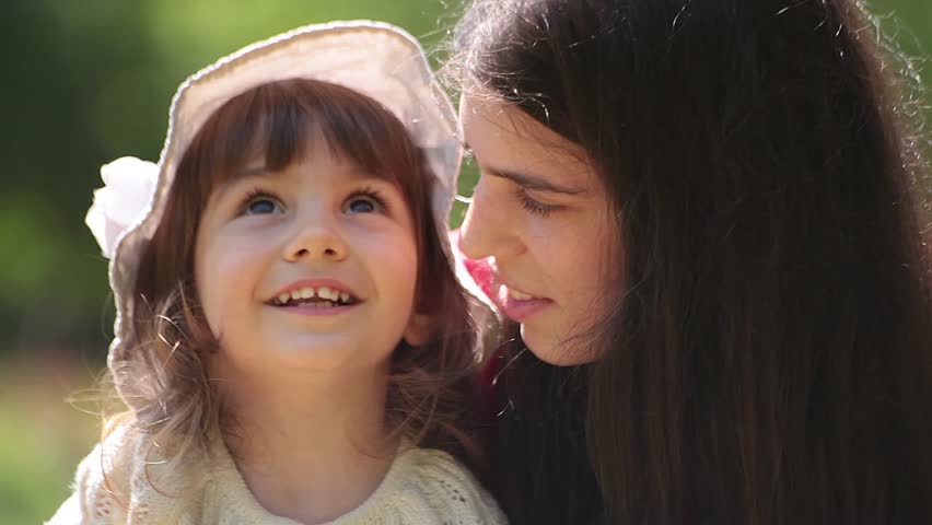 Brunette young adult mother talks to her little baby girl. Sunny day, outdoor portrait.
