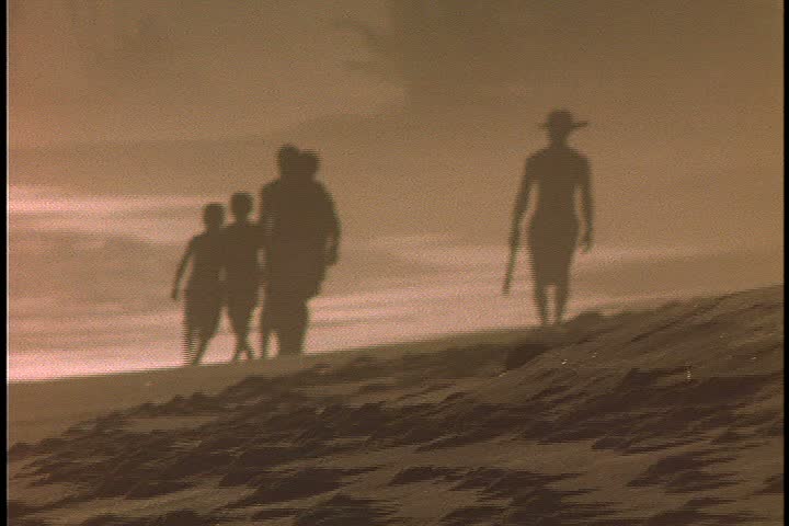 Slavery reenactment in Ghana, Africa. WS of silhouettes of an overseer leading a group of five chained men down the beach. Overseer raises a big stick to strike one of them.