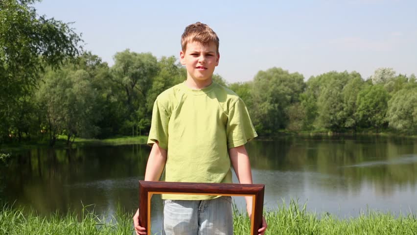 Boy in green t-shirt raises picture frame to level of head, near pond at summer park