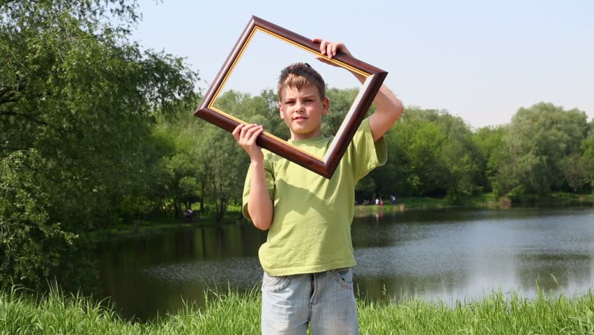 Young boy holds picture frame and rotates it near near his face, on bank of pond in park at summer sunny day