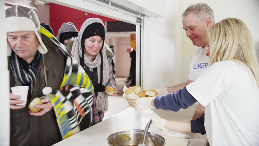 Friendly voluntary workers standing at a soup kitchen serving hatch are handing out cups of hot soup and bread to a waiting line of homeless and needy people. 