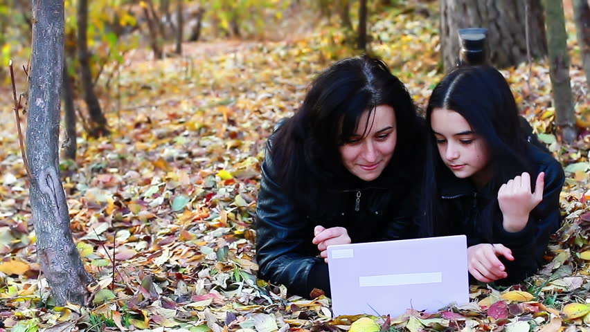 Young woman and teenager laying on leaves with laptop. Young mother and daughter working on computer. 