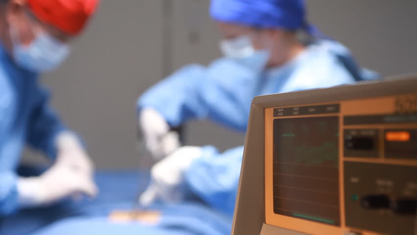 Focus on a heart rate monitor with two surgeons working in the background.