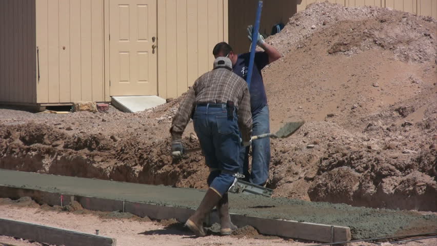 Water is sprayed while a man shovels more cement into the forms. Then another man uses a bull float to smooth the cement.