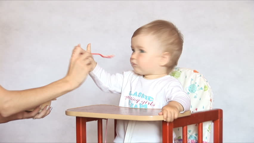 Baby sits at a children's little table. Mum feeds the child with porridge. Isolated on the white