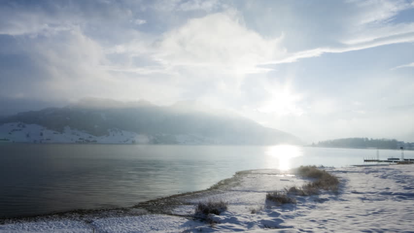 A living winter postcard: Lake, sun reflections, and snow covered hills (time lapse)
