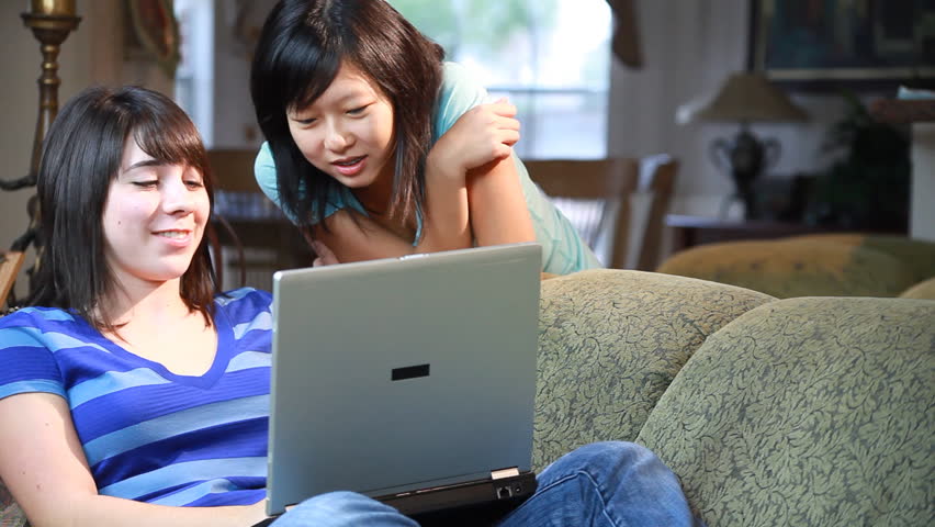 A young Asian teenager and her friend, relaxing on a couch, talking and using a laptop.