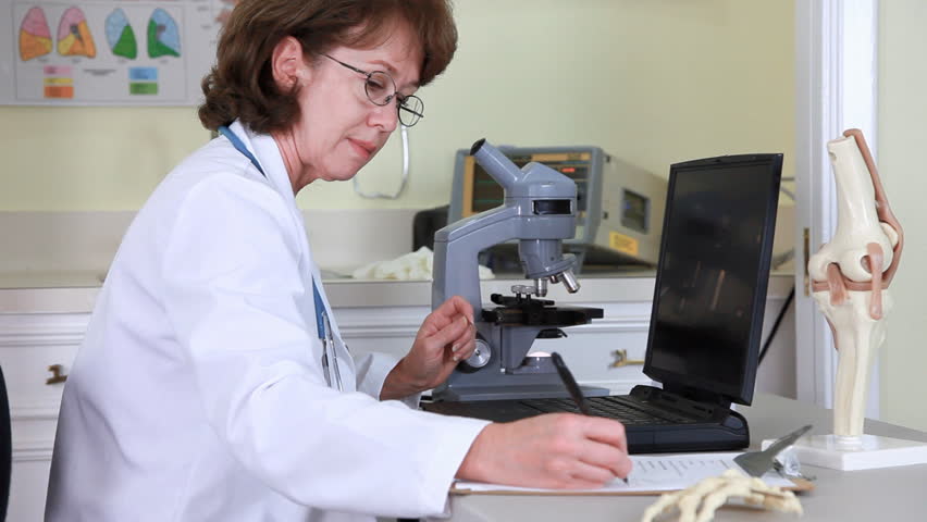 A female physician looking through a microscope using a laptop to record her findings.