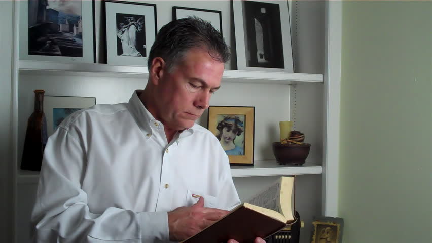 A man in front of a bookcase leisurely reading a book, answers his cell phone and begins to talk.