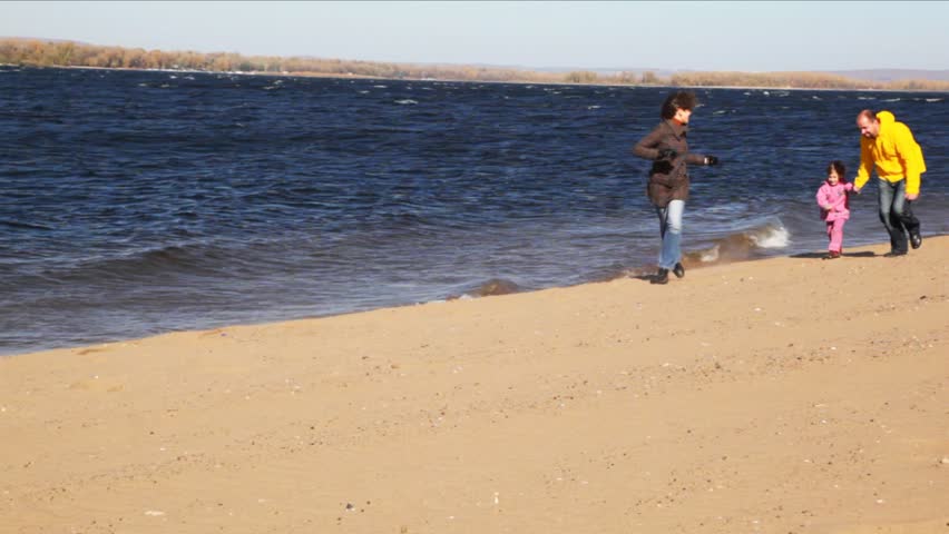 family of mum, daddies and daughter runs one after another on sandy to river bank 