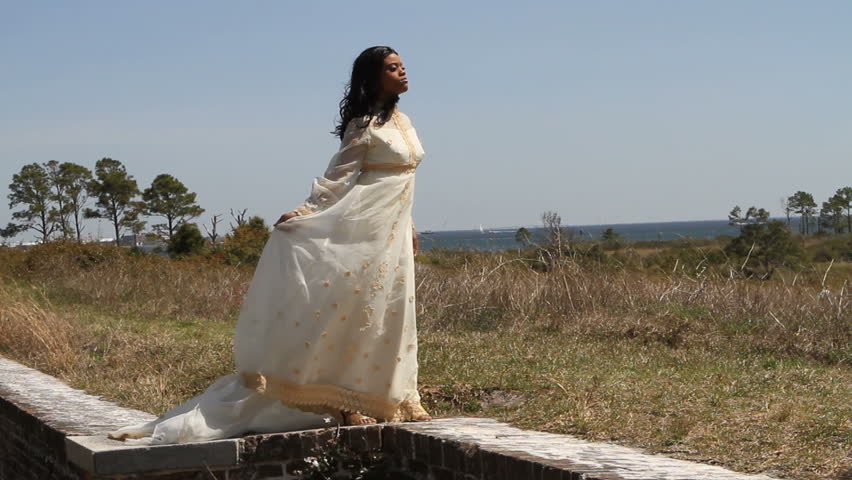 Black woman in a 1960's era bridal gown stands on a brick wall with the wind blowing across her dress.