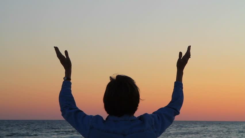 Mature woman raises her hands while praying at the seashore.