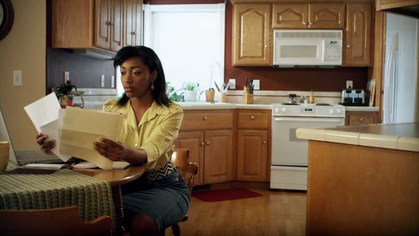 An African American woman performs various tasks around the house. She searches for a job, pays bills, makes coffee, studies the bible, reads a book, and works on her laptop.
