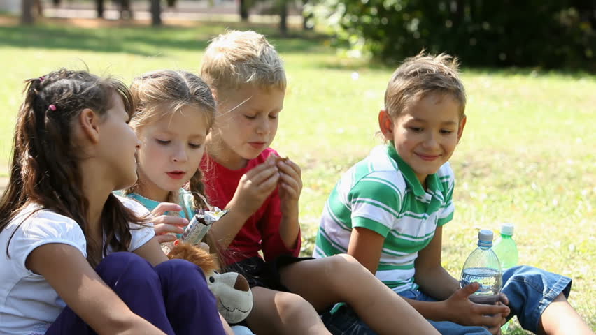 Five children sitting on grass in the park and eating, one girl drinking mineral water 