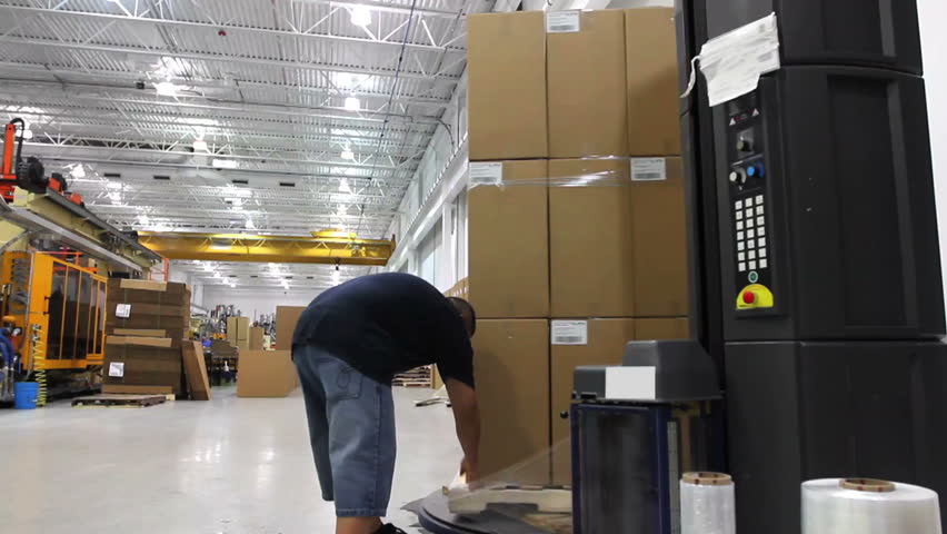 A worker starts a shrink wrap machine on a large stack of boxes in a factory. 