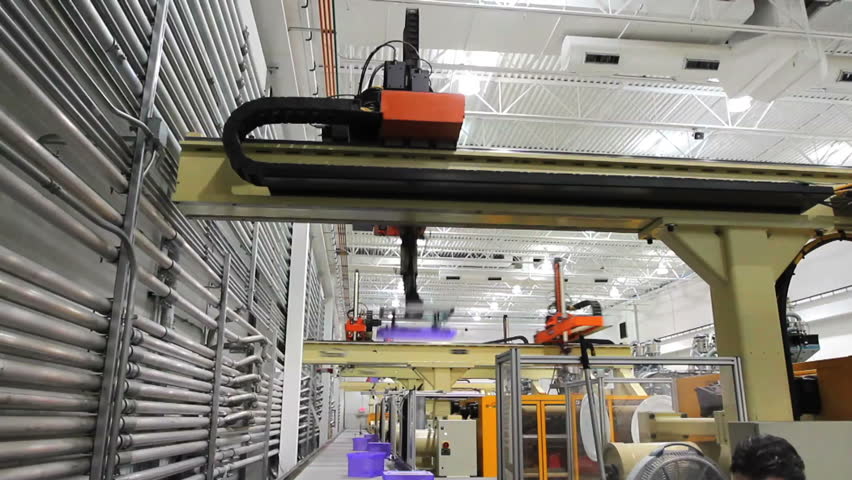 A factory supervisor inspects products as they come down a conveyor belt with robotic arms moving in background. 