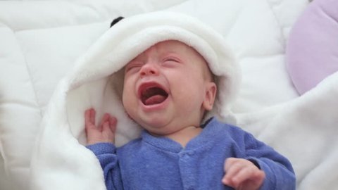Newborn crying baby boy. New born child tired and hungry in bed under a blue knitted blanket.