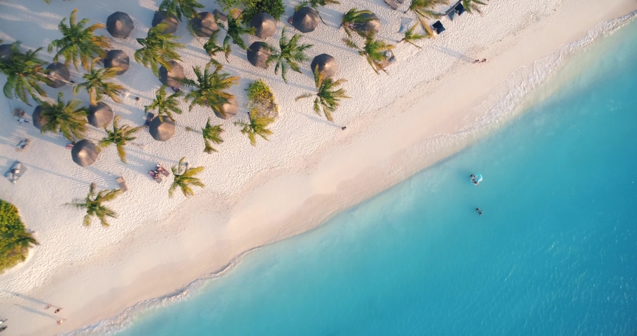 Aerial view of sea waves, umbrellas, green palms on the sandy beach at sunset. Summer in Zanzibar, Africa. Tropical landscape with palm trees, people, parasols, sand, blue water. Top view from air