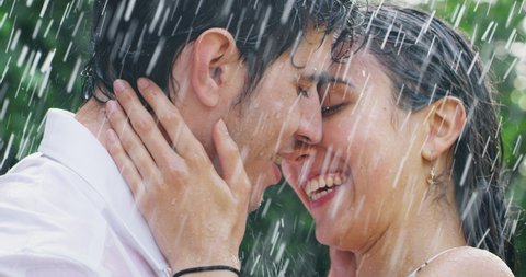 Slow motion of young carefree couple in love are hugging and kissing under  the rain on a background of green trees.