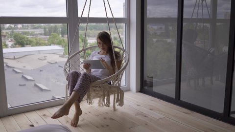 Pretty Young Woman Swinging In A Hanging Chair While Using Her Tablet At The Large Floor To Ceiling Window Freelance Concept The Woman Relaxing At Home