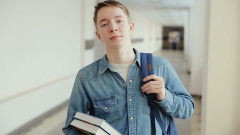 Portrait of a happy male caucasian student standing in a busy hallway with books and backpack