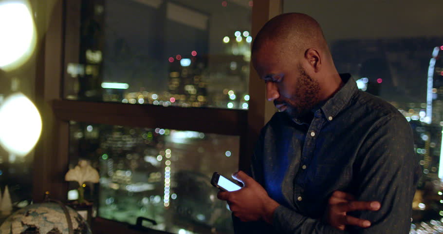 Handsome African American man using a smart phone (graphics removed) in Downtown Los Angeles apartment at night.  Locked off mid shot with with city buildings in background.