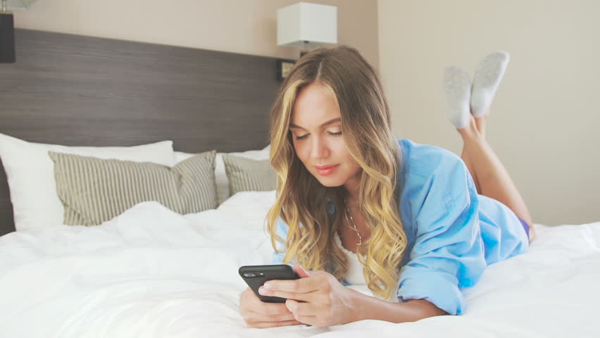 Beautiful Woman Lies On A Bed On Stomach Communicates On Mobile Phon