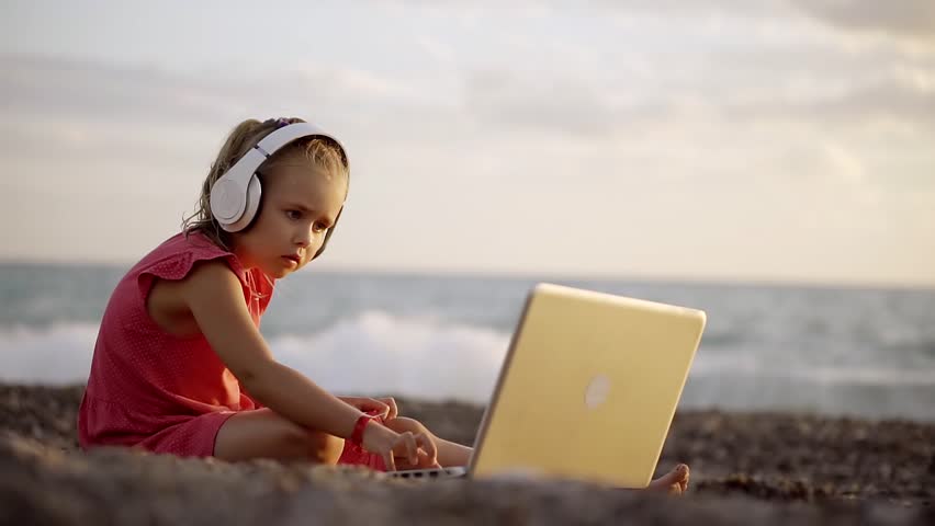 Little Girl Is Sitting On The Beach Near The Sea And Is Watching A Cartoon On Little Girl Is Sitting On The Beach Near The Sea And Is Watching A Cartoon On